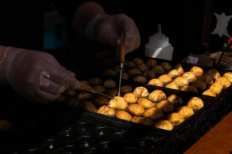 Hands turning takoyaki balls on a hot griddle at a Japanese street food stall.